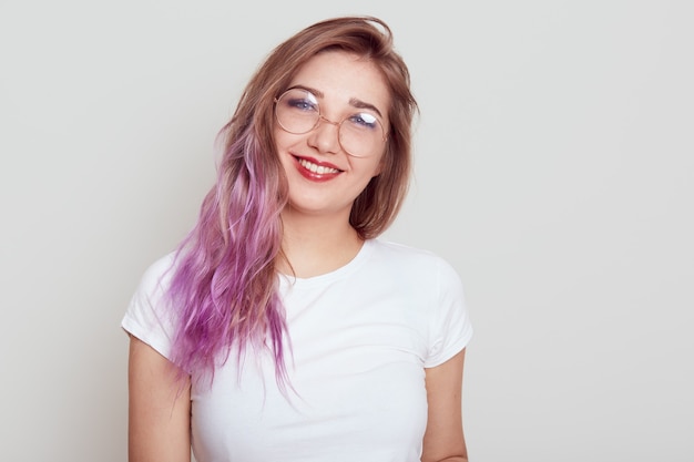 眼鏡をかけた心地よい女性と、カメラに向かって直接笑顔を見せている白いカジュアルなTシャツは、灰色の背景の上に孤立した、前向きな感情を表現する薄紫色の髪をしています。