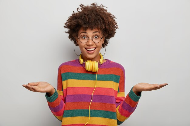 pleasant looking female raises both palms, holds copy space, has friendly expression, wears glasses, casual sweater, looks directly at camera, uses headphones