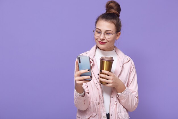 Pleasant looking female wearing spectacles and pale rose jacket posing isolated over lilac wall, drinking hot beverage from thermo mug, holding smart phone in hands and browsing Internet.