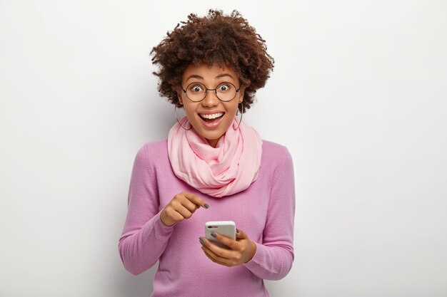 Pleasant looking female with crisp hair, points at cell phone device, donwloads new modern application, has happy expression, wears spectacles for vision correction, purple jumper and silk scarf
