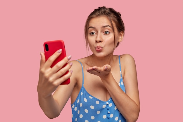 Pleasant looking girlfriend flirts with man over smart phone, makes air kiss during video call, holds mobile phone in front, wears casual summer dress