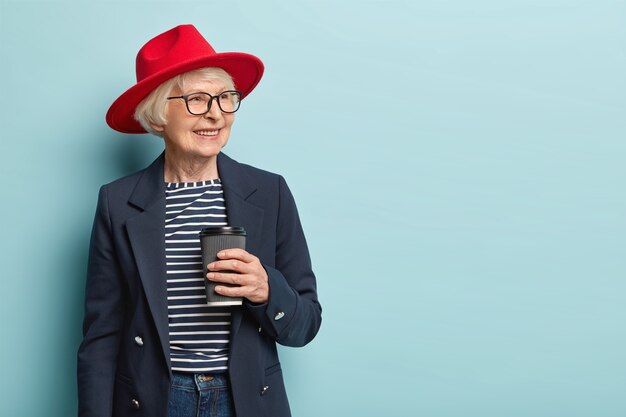 pleasant looking glad mature woman expresses positive emotions, wears elegant jacket and red hat, holds disposable cup of coffee, drinks hot beverage, focused aside, smiles broadly