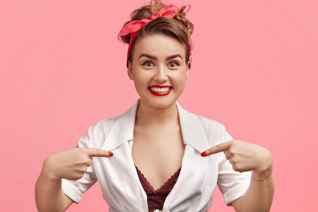 Pleasant looking happy young woman wears stylish red headband and white shirt, points with both index fingers at herself