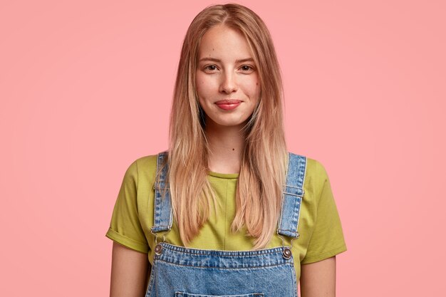 pleasant looking light haired woman with satisfied expression, dressed in fashionable overalls, looks joyfully