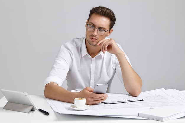 Pleasant looking male architect looks seriously into tablet computer, works with notes and sketches, drinks coffee, being very busy. Talented young male engineer works on construction project