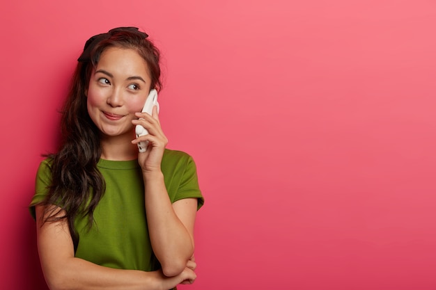 La ragazza bruna naturale dall'aspetto piacevole tiene lo smartphone vicino all'orecchio, gode di una bella conversazione telefonica, guarda da parte, indossa una maglietta casual, discute qualcosa di interessante con un amico, isolato sul muro rosa