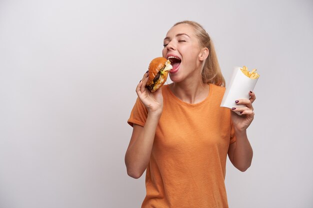 Free photo pleasant looking positive young blonde lady with ponytail hairstyle keeping eyes closed while enjoying her tasty burger, standing over white background in casual clothes