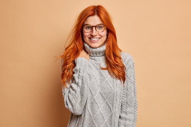pleasant looking red haired young woman smiles broadly hears something very pleasant has friendly talk with best friend wears warm winter sweater.