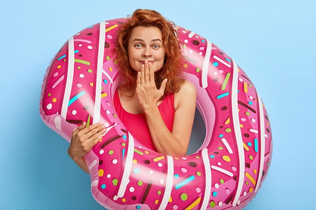 Pleasant looking redhead younng woman giggles happily, keeps hand on mouth, satisfied with amazing view of sea, dressed in red bikini, carries big rubber swimring, has summer rest