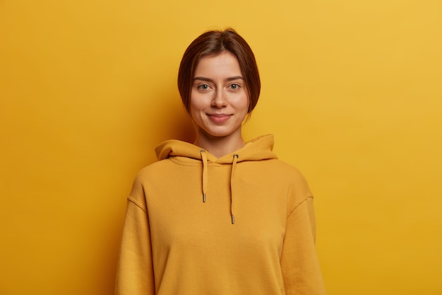 Pleasant looking teenage girl wears comfortable hoodie, had combed dark hair, looks at camera with little smile