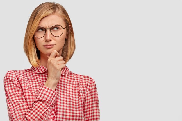 Pleasant looking thoughtful woman tries to solve problem, keeps hand under chin