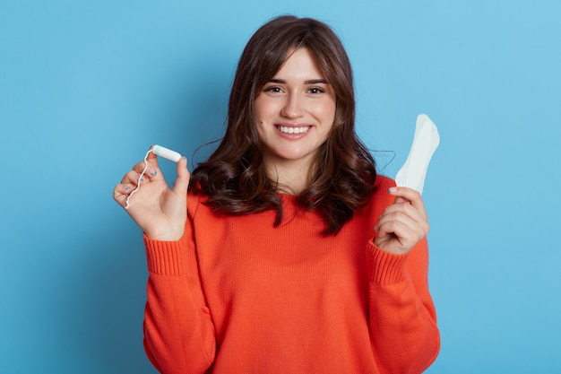 Pleasant looking woman in casual attire standing with tampon and sanitary napkin in hands