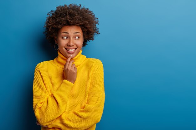 Pleasant looking woman with Afro hair looks aside with chuckle, shows white teeth, dressed in yellow jumper, caught eye of handsome guy, isolated over blue background