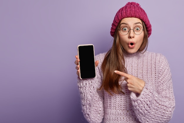 Pleasant looking woman with shocked expression, points at empty screen of cell phone, shows free space, demonstrates modern electronic device, wears winter clothes, isolated against purple wall