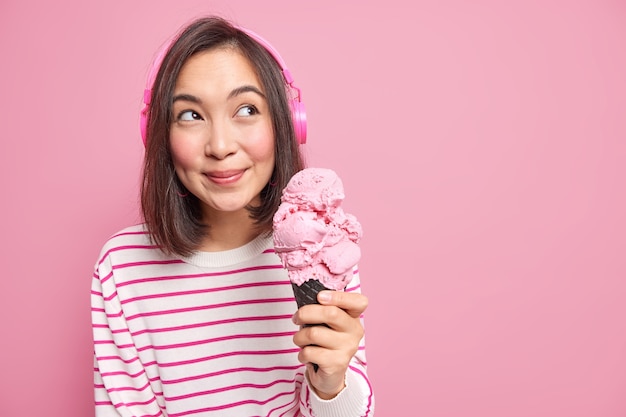 pleasant looking young Asian woman with dark hair holds big strawberry ice cream looks away thoughtfully wears stereo headphones isolated over pink wall copy space area.