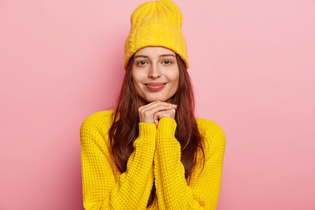 見栄えの良い若い黒髪の白人女性は、優しい笑顔を浮かべ、あごの下に手を保ち、青い目をして、スタイリッシュな黄色の帽子とニットのセーターを着て、バラ色のスタジオの壁を越えてモデルを作ります