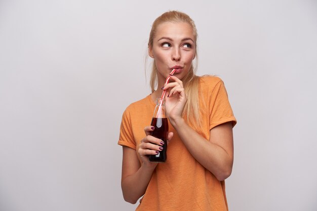 Pleasant looking young positive long haired blonde woman with casual hairstyle drinking soda with straw from glass bottle, dressed in orange t-shirt while standing over white background