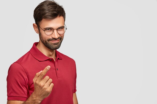 Pleasant looking young unshaven guy with mysterious look, has pleased expression, intention to do something, wears casual red t-shirt, isolated over white wall with blank space for text