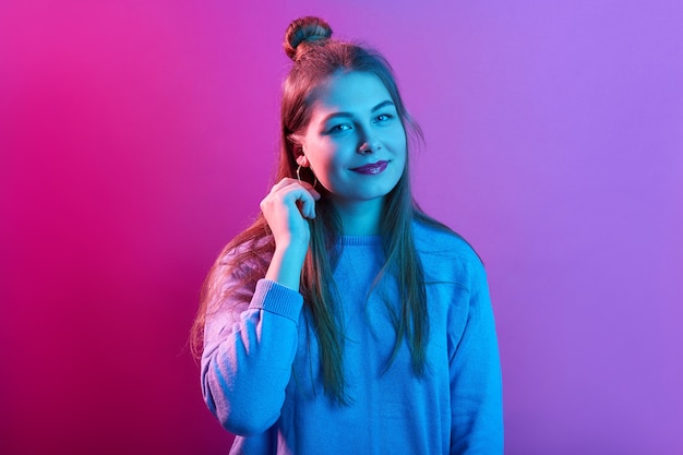 Pleasant looking young woman wears casual sweater, has hair bun, looks with calm facial expressions, poses against pink neon space