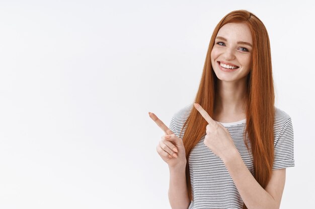 Pleasant pretty redhead young girl blue eyes wearing summer striped t-shirt pointing upper left corner index fingers presenting awesome product advertisement smiling happily, white wall