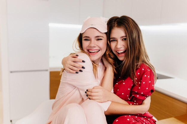 Pleasant sisters with shiny hair embracing in morning. Cute european lady in stylish sleepmask laughing to camera.