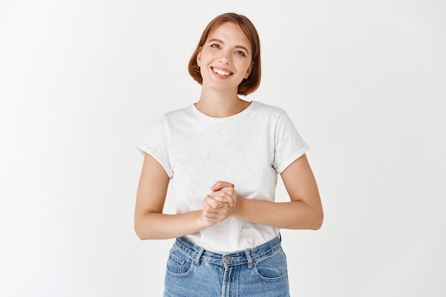 Pleasant smiling girl looking friendly, holding hands together, consultant ready to help, standing on white wall