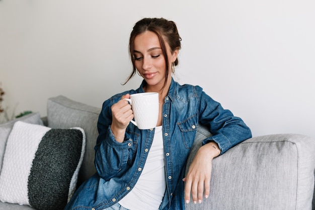 Pleasant stylish girl with tanned skin and dark collected hair is wearing denim shirt is holding coffee and resting at home