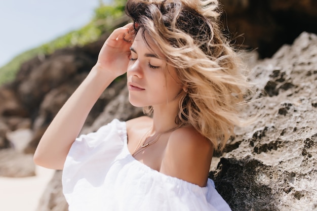 Piacevole donna in posa vicino a roccia con gli occhi chiusi. ritratto all'aperto di signora sensuale abbronzata in piedi sulla natura.