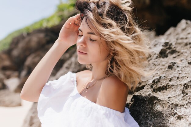 Pleasant woman posing near rock with eyes closed. Outdoor portrait of tanned sensual lady standing on nature.