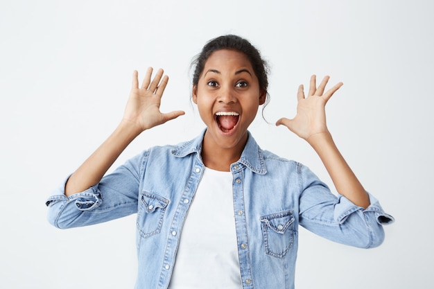 Pleasantly surprised Afro-american woman looking with bugged eyes holding her hands up, opening her mouth in amazement not expecting to recieve such great present. Happy dark-skinned model isolated ov