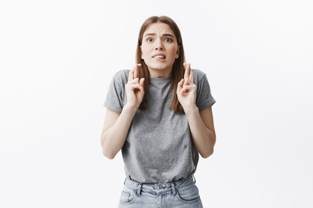 Please. Close up of handsome young dark-haired female student in casual grey clothes crossing fingers, biting lips,  with frightened expression, waiting for test results