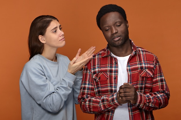 Free photo please forgive me. unhappy worried young caucasian woman gesturing having mournful facial expression, asking her offended upset dark skinned husband for forgivenes. people and relationships