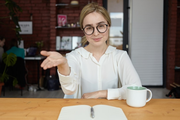 Please, have a seat. Indoor shot of beautiful friendly looking young Caucasian businesswoman in eyewear smiling and making welcome gesture during business meeting with partner or client at cafe