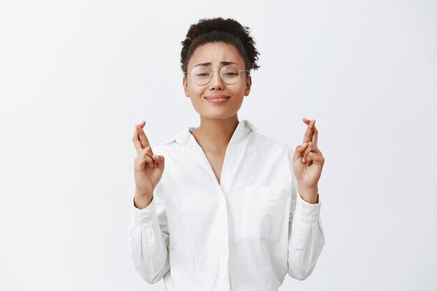 Please help me out, beg you. Charming and cute dark-skinned woman in glasses and white-collar shirt, holding fingers crossed and smiling tight while praying and begging for lending money from friend