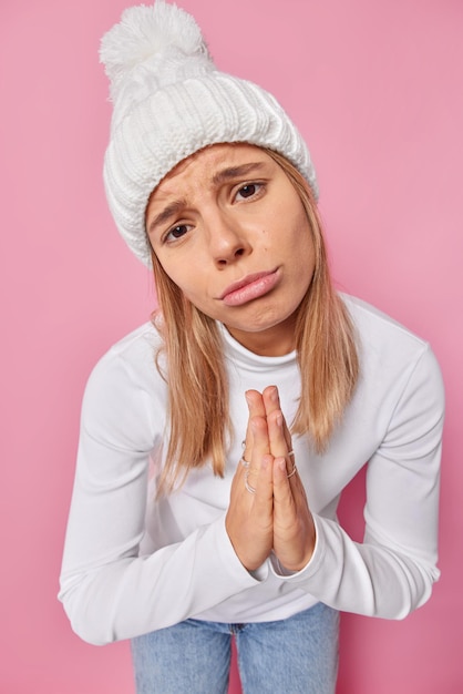 Please help me. Sulking upset young woman presses palms together looks with begging expression as asks for favor needs advice wears knitted hat and casual jumper isolated over pink background