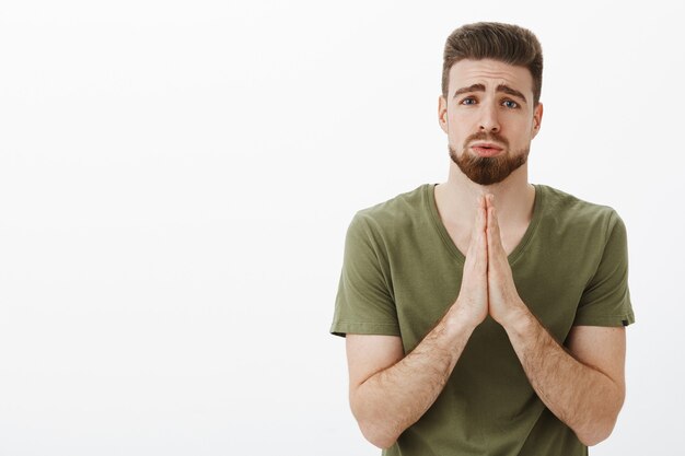 Please I beg you. Portrait of cute charming bearded male in need holding hands in pray, supplicating pouting and frowning looking upset as asking favour or help hopefully over white wall