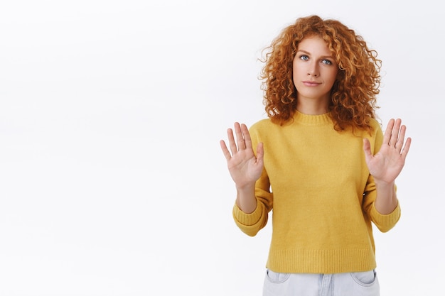 Per favore, fermati, stai lontano da me. la donna riccia rossa assertiva sa cosa vuole, alza le braccia in segno di divieto, movimento di rifiuto, disapprovazione scuotere la testa, sorridere, partecipazione riluttante, muro bianco