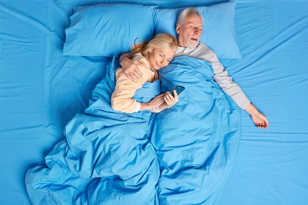 현대 기술에 중독 된 여성은 침대에서 휴대 전화를 사용하여 잠자는 남편의 포옹을 받아 잠들기 전에 인터넷을 스크롤합니다. 중년 가족 부부는 아늑한 침실에서 휴식을 취합니다.