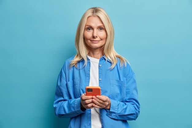 Pleased adorable blonde middle aged woman holds smartphone uses mobile phone