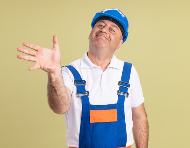 Pleased adult builder man in uniform holding hand open on olive green