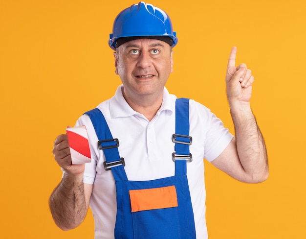 Pleased adult builder man in uniform holds covers mouth with duct tape and points up isolated on orange wall