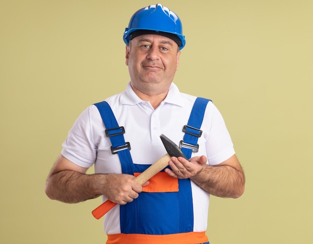 Pleased adult builder man in uniform holds hammer isolated on olive green wall