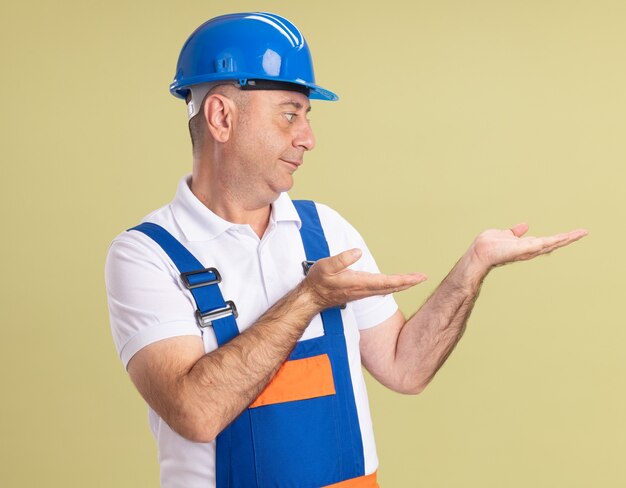 Pleased adult builder man in uniform looks at empty hands isolated on olive green wall