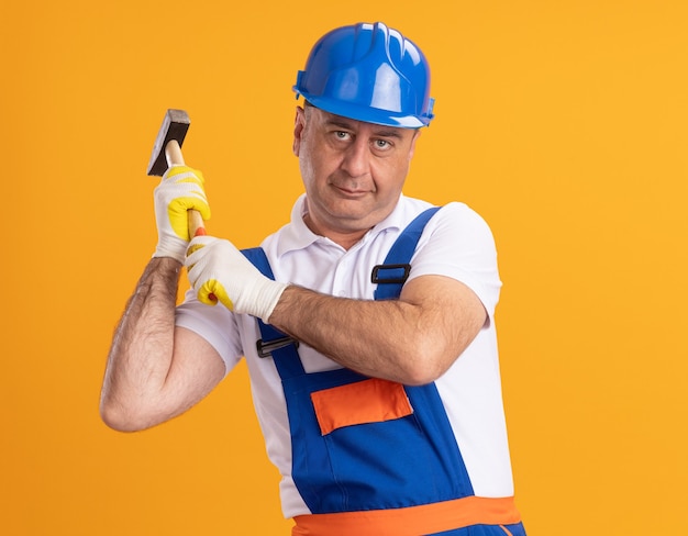 Pleased adult builder man in uniform wearing protective gloves holding hammer isolated on orange wall