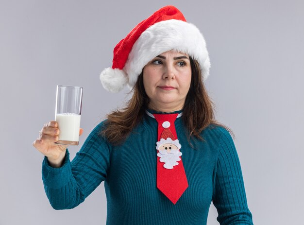 Pleased adult caucasian woman with santa hat and santa tie holding glass of milk looking at side isolated on white wall with copy space