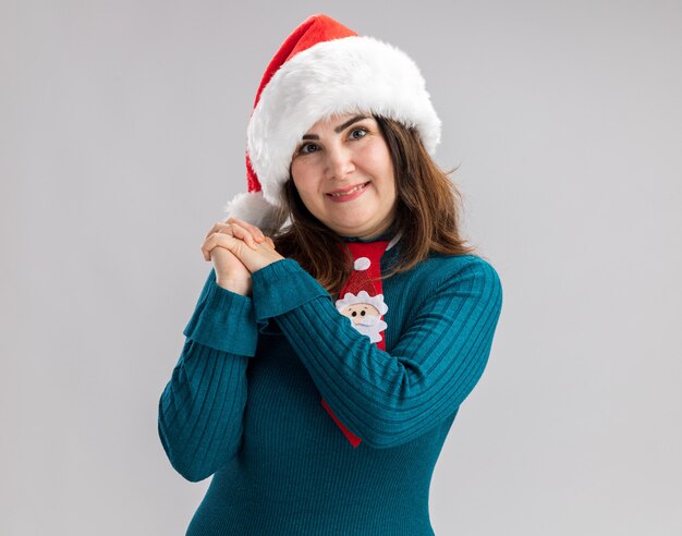 Pleased adult caucasian woman with santa hat and santa tie holding hands together isolated on white wall with copy space