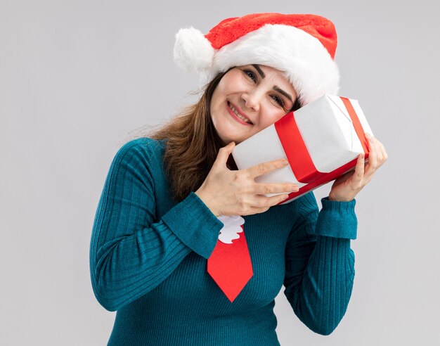 サンタの帽子とサンタのネクタイを保持し、コピースペースと白い背景で隔離のクリスマスギフトボックスに頭を置くと大人の白人女性を喜ばせる