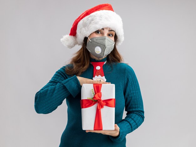 Pleased adult caucasian woman with santa hat and santa tie wearing medical mask holding christmas gift box isolated on white wall with copy space