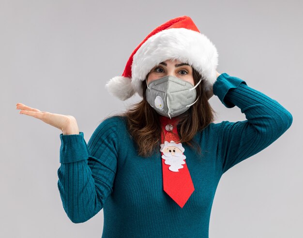 Pleased adult caucasian woman with santa hat and santa tie wearing medical mask puts hand on head and keeps hand open isolated on white background with copy space