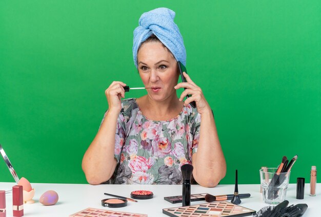 Pleased adult caucasian woman with wrapped hair in towel sitting at table with makeup tools talking on phone applying lip gloss 